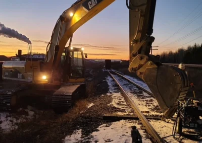 A CAT excavator works beside railway tracks at sunset, with smoke rising in the background and snow covering the ground.