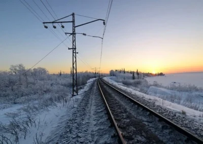 Snow-covered railway tracks curve through a winter landscape, framed by frosted trees and a vibrant sunset on the horizon.