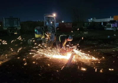 A worker in safety gear sparks fly while cutting metal at night, illuminated by bright lights against a dark, industrial background.