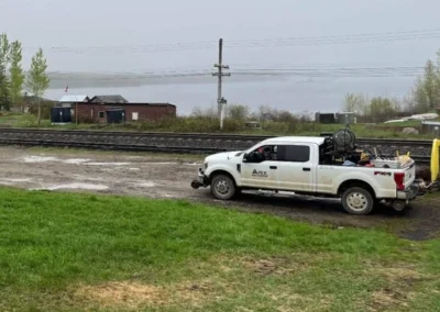 A white pickup truck is parked near train tracks, with a cloudy sky and green grass in the foreground; a rustic building is in the background.