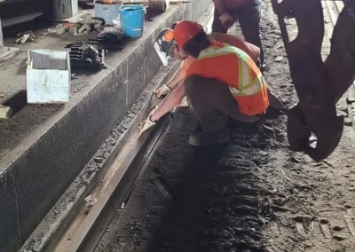 Two workers in orange safety vests and helmets inspect a track, with tools and equipment scattered on the ground in a dim workspace.