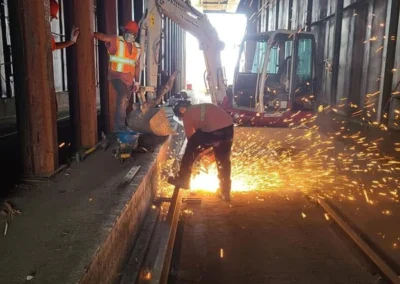 Construction workers in safety gear cutting metal, creating sparks, alongside an excavator in a semi-enclosed industrial space.